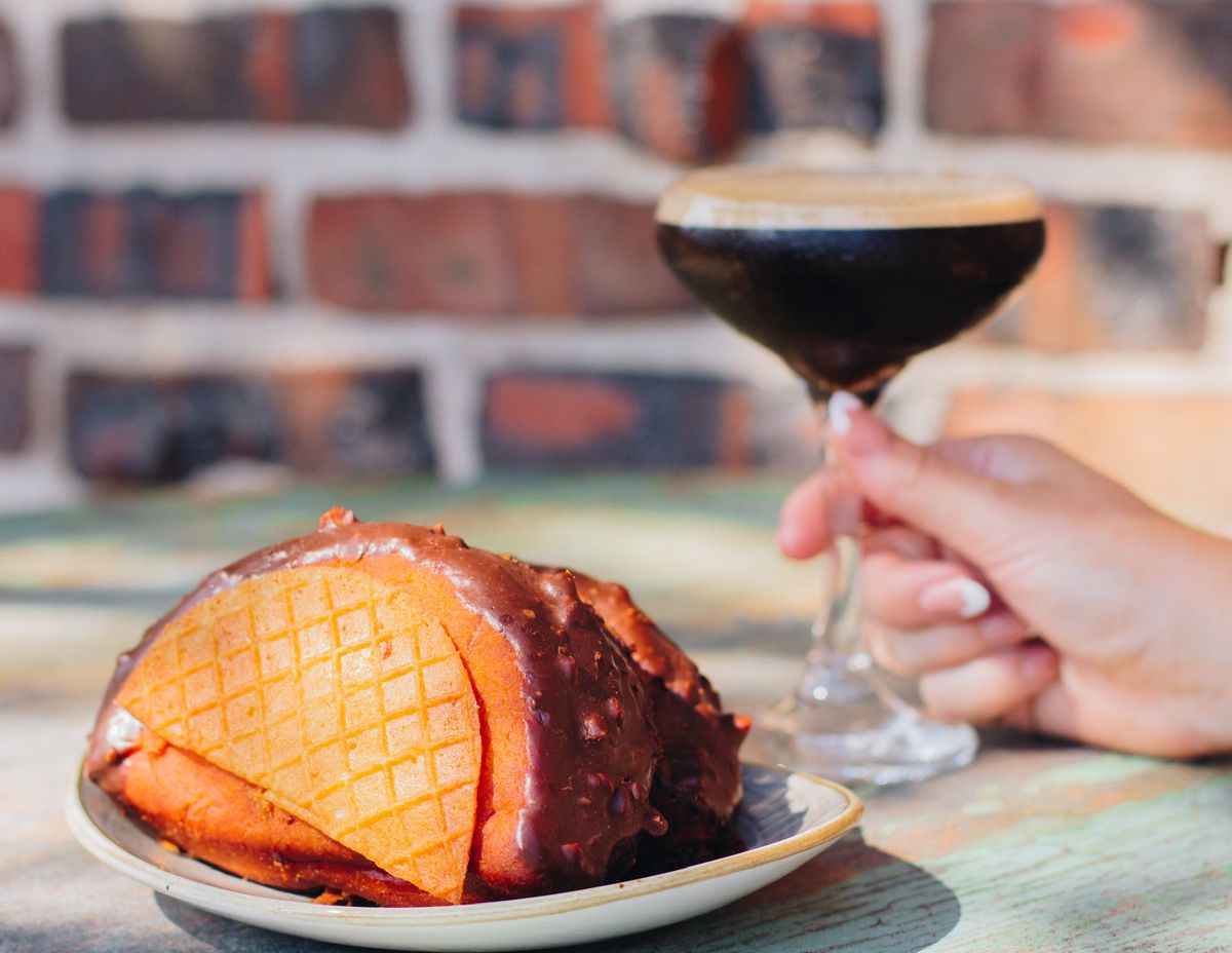 A half-moon shaped donut has chocolate and waffle cone fillings, making it look like a Choco Taco.  A hand holds a dark brown cocktail in the background.